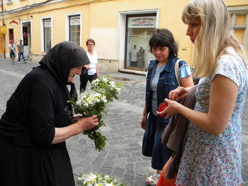 Două doamne se apropie de bătrânica din colțul străzii, care vinde flori! Cu o mână tremurândă, bătrâna apucă 2 buchete de flori, ridică privirea spre cea din fața ei, și îi spune…